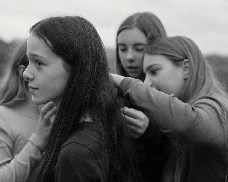 Three young women are outdoors in a black and white portrait. One is getting her hair styled by the other two, who are standing behind her.