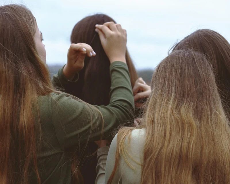 Four women stand close outdoors with light hair. One woman adjusts another woman's hair, viewed from behind in soft, natural light.