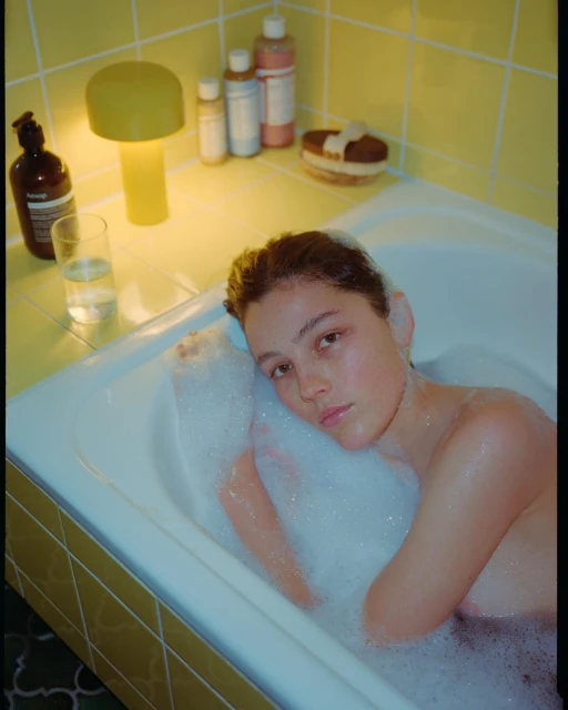 Woman soaks in a bubble bath in a yellow-tiled bathroom. The scene includes bath products, a glass of water, and a yellow lamp.