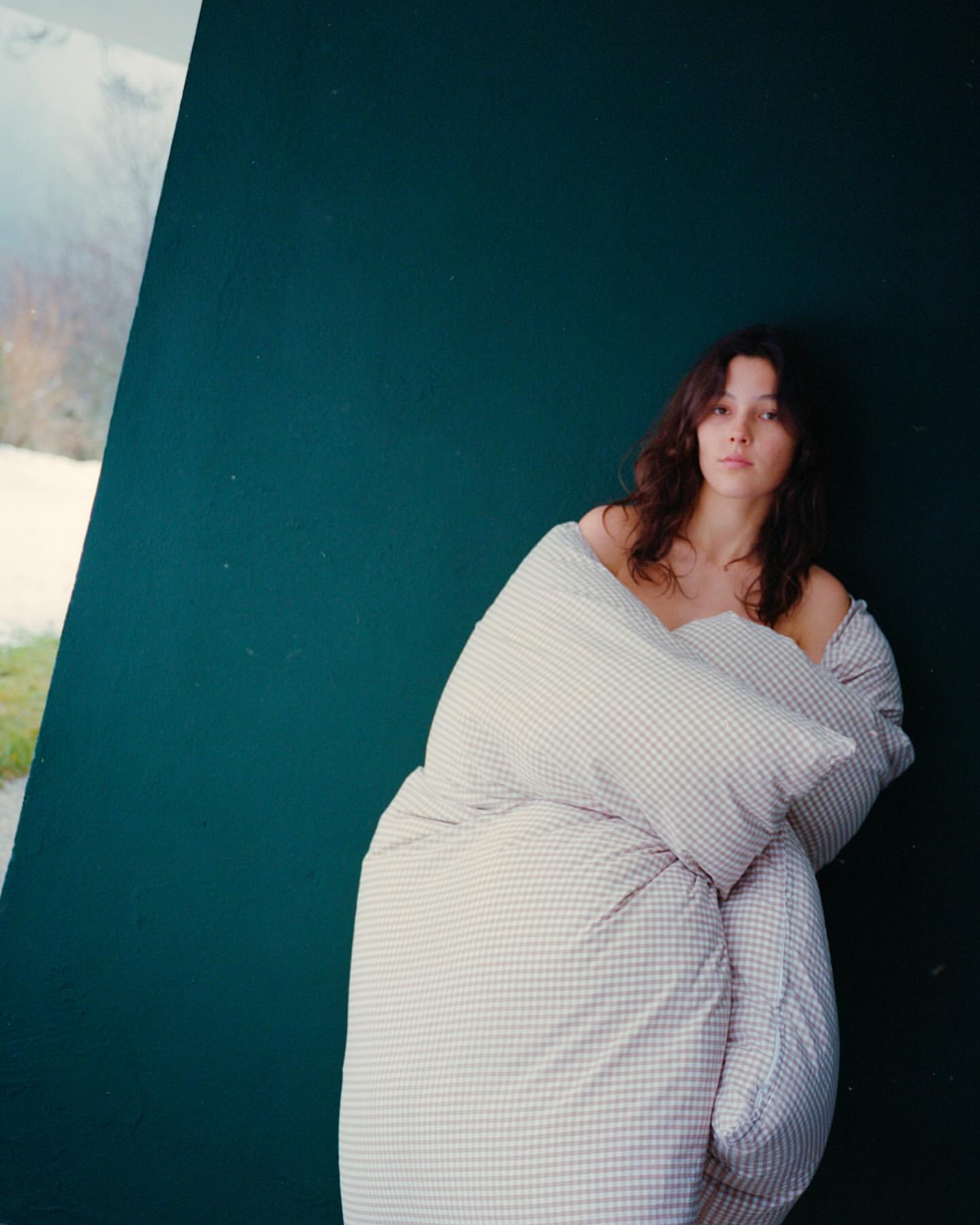 Portrait of a young adult woman wrapped in a checkered duvet, posed against a teal wall with natural outdoor lighting.