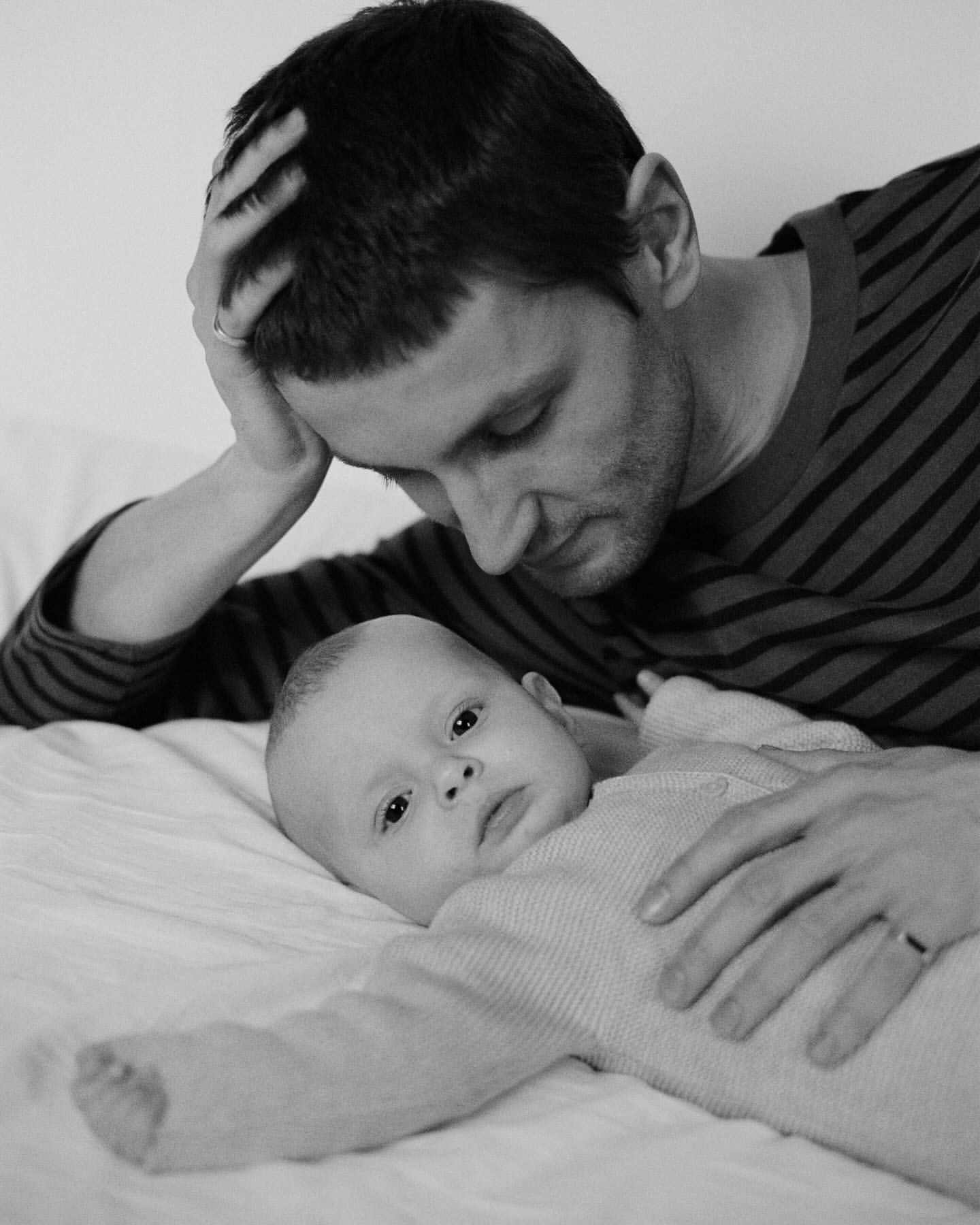 Black and white image of a father lying next to his baby on a bed. The baby looks at the camera, while the father looks down at the infant.