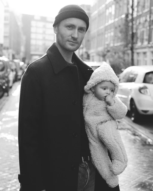 Black and white shot of an adult holding a baby in an urban environment. The background shows buildings, parked cars, and street details.