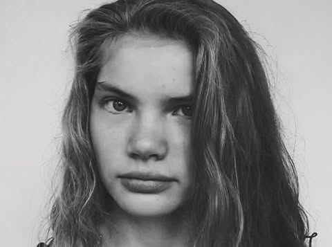 Close-up black and white portrait of a young female model with long dark hair looking directly at the camera.