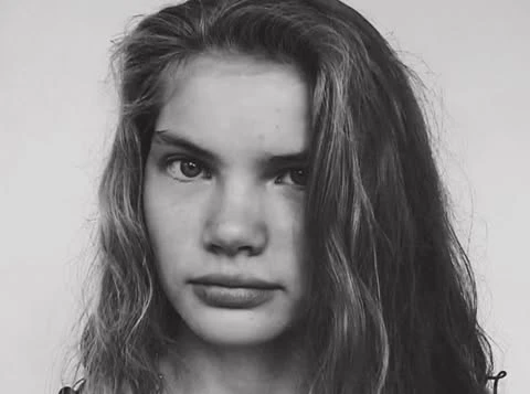 Close-up black and white portrait of a young female model with long dark hair looking directly at the camera.