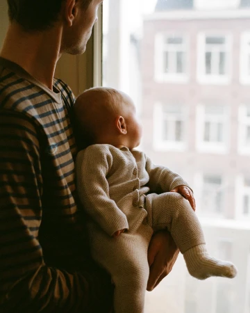 Man holding a baby in a knitted onesie while looking out a window at a city street