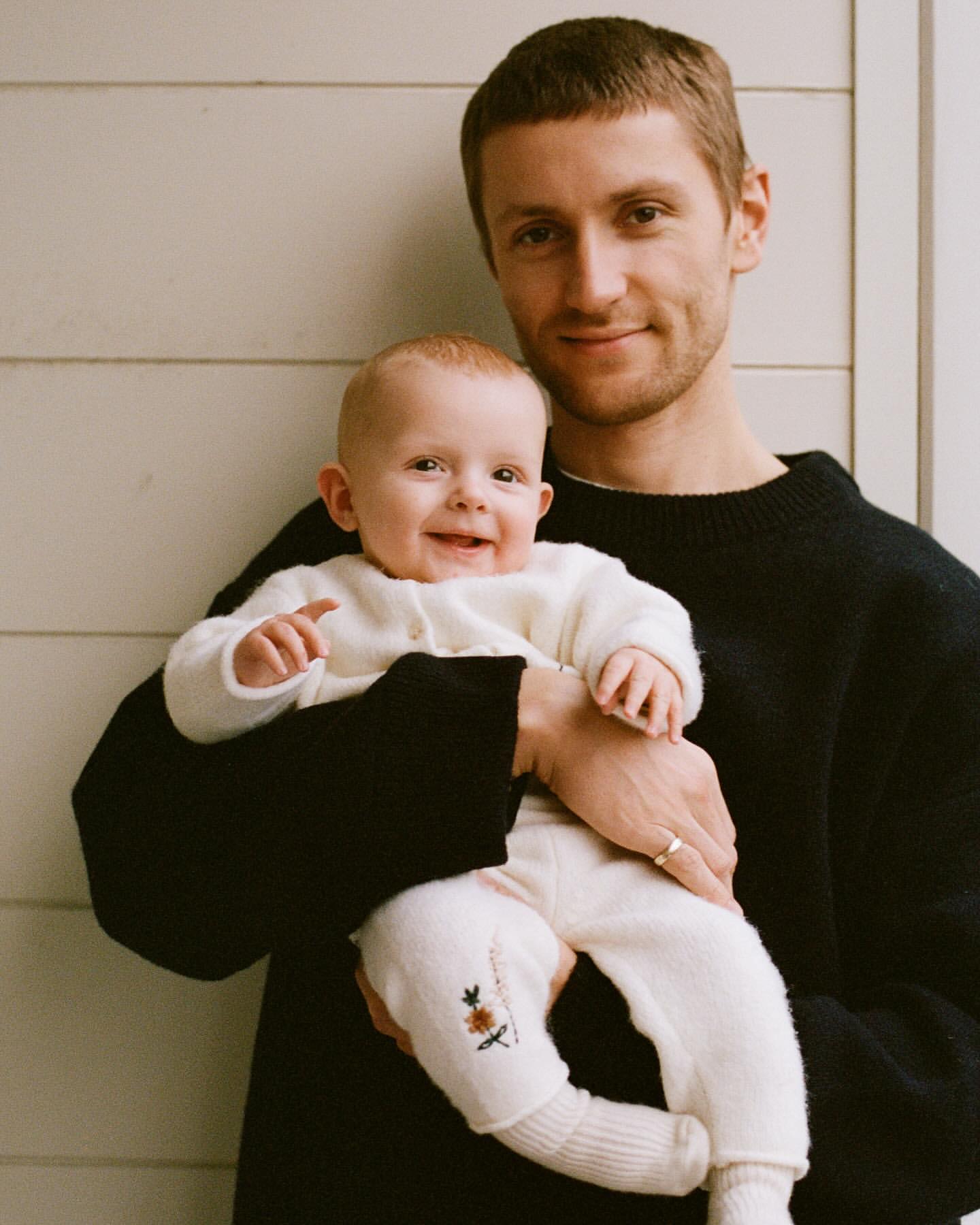 A young man in a black sweater holds a baby in a white outfit, both smiling in a bright indoor setting with a neutral-toned plank wall background.