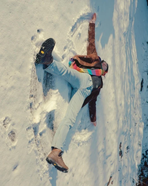 Aerial shot of a person lying in snow, creating a snow angel, wearing a brown jacket, light blue jeans, and brown boots.