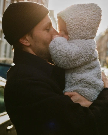 Man wearing black beanie and coat holding a baby in a grey hooded fleece onesie outdoors