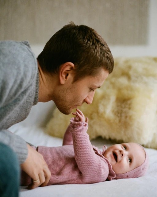 Father and baby daughter interact on bed. The baby wears a pink knit outfit and lies on a white bedsheet as dad looks on.