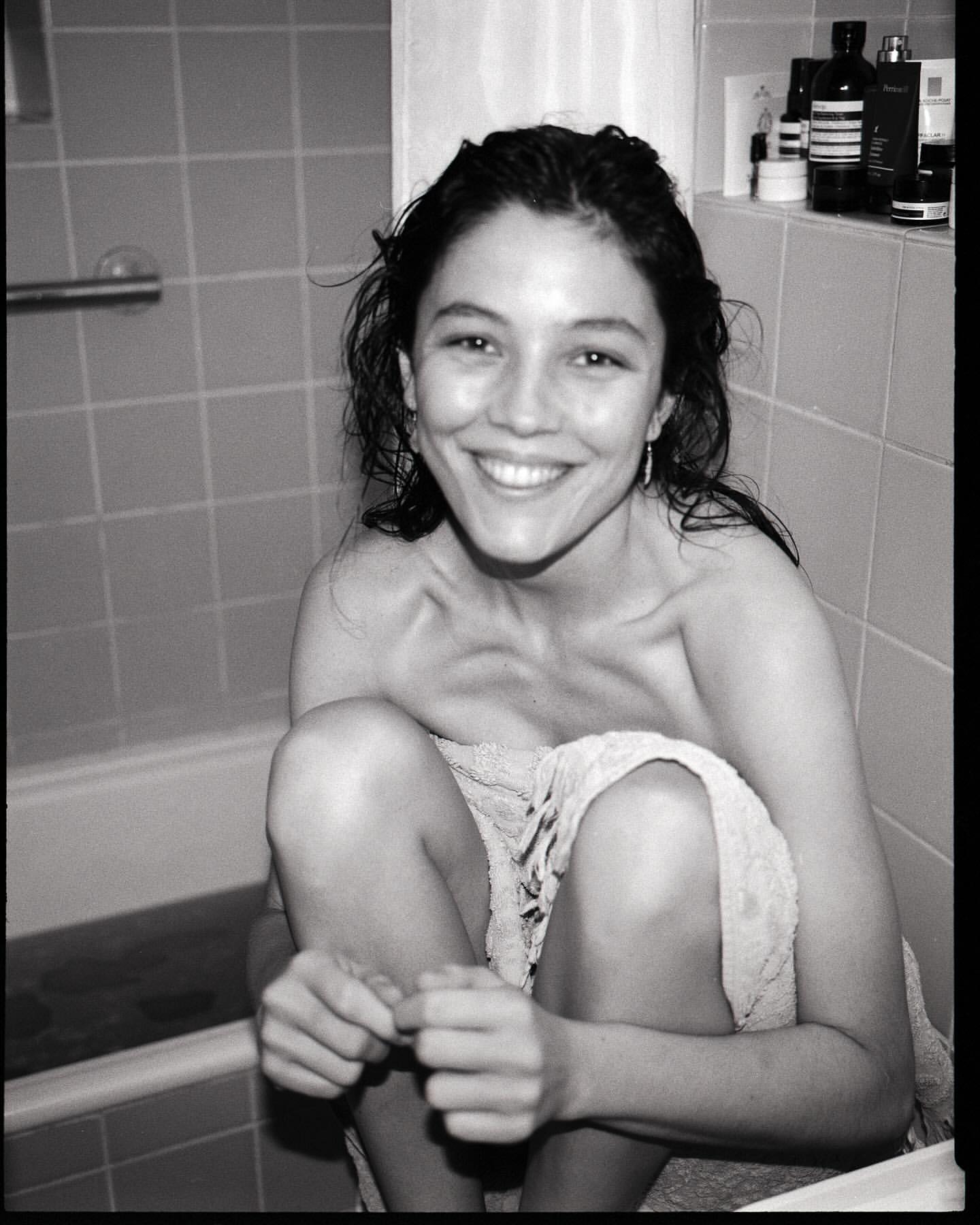 Black and white portrait of a smiling woman sitting in a tiled bathtub, wrapped in a towel, with assorted bottles on a shelf behind her.