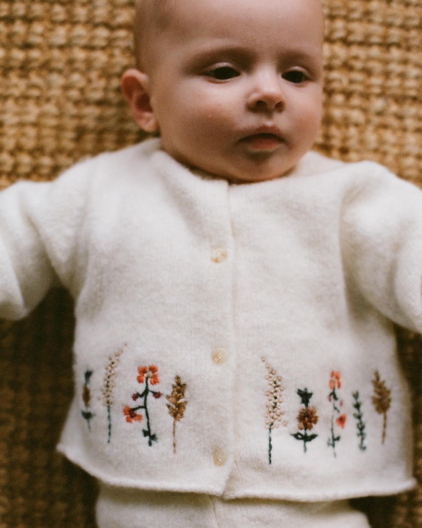 Close-up of a baby lying on a woven surface, wearing a cream-colored cardigan with delicate floral embroidery, looking at the camera.