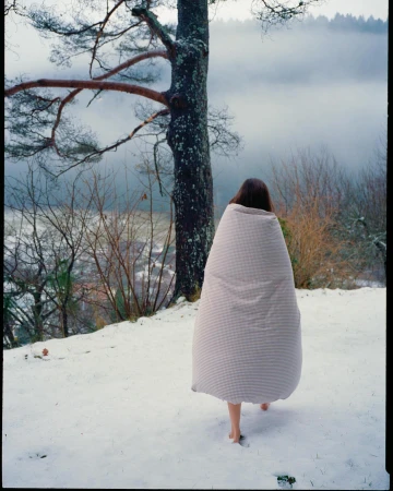 A woman covered in a blanket walks barefoot in a snowy outdoor setting with a tree and fog-covered landscape in the background.
