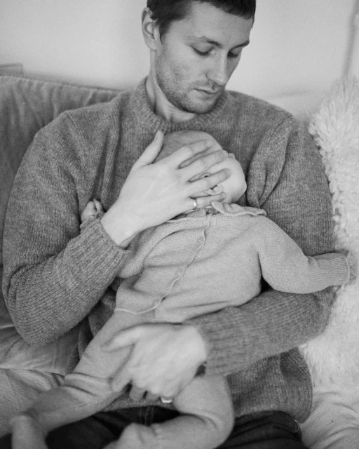 Monochrome portrait of an adult male holding a baby. The man wears a crewneck sweater, and the baby is in a knitted cardigan.