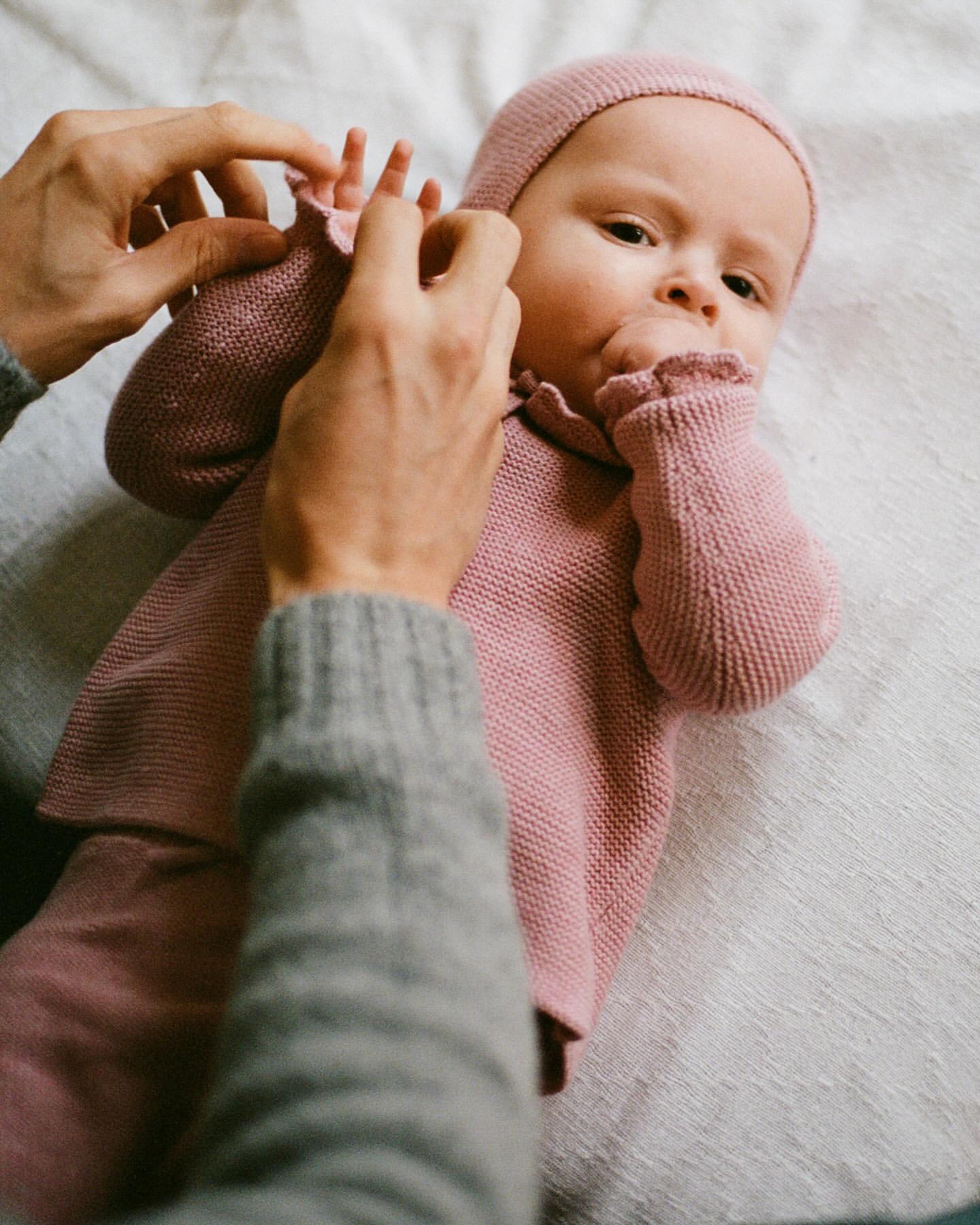Infant in a pink knit outfit lying on a textured surface, attended by an adult adjusting their sleeve; warm, soft lighting.