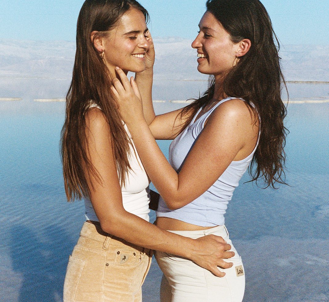 Two adult women in light-colored tops and bottoms pose closely on a beach with a light blue sea and sky creating a warm, intimate mood.
