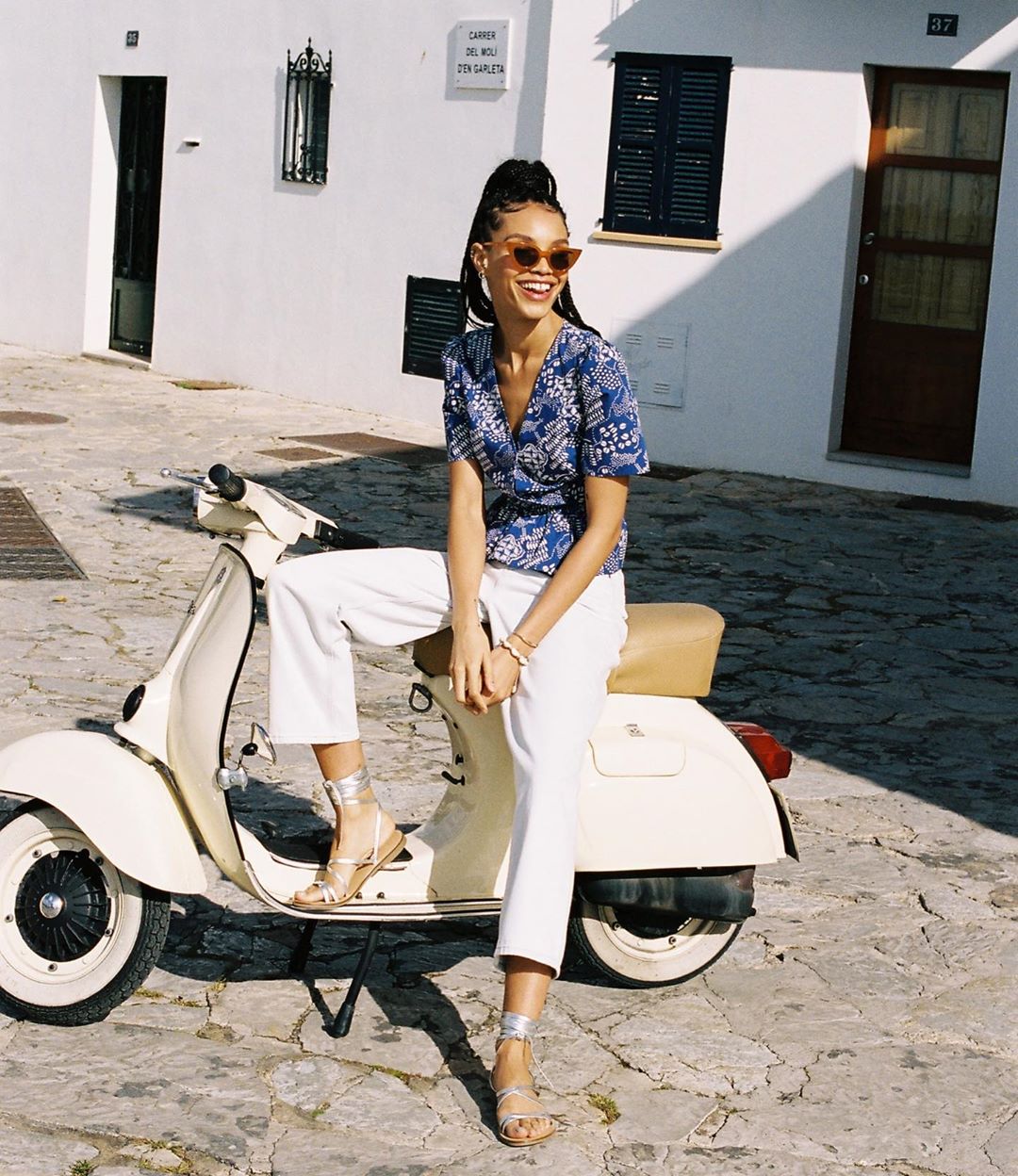 Smiling woman sits on a vintage scooter in an outdoor urban setting, wearing a blue wrap top, white pants, and strappy sandals.