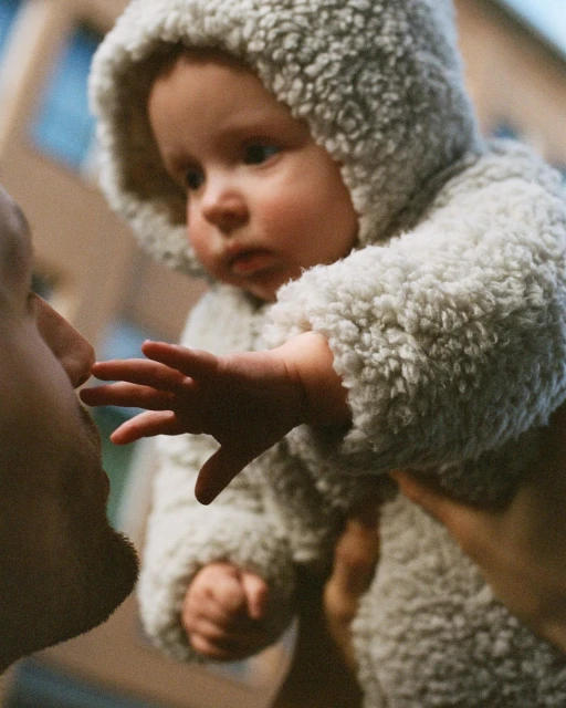 Adorable baby in a hooded outfit reaching towards an adult's face, capturing a heartwarming moment, soft focus, natural light.