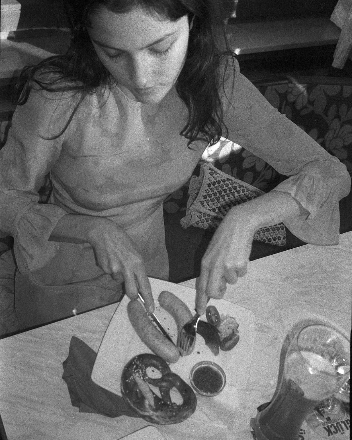 Monochrome image of a young woman at a table indoors, cutting into sausages with a fork and knife next to a pretzel and beverage.