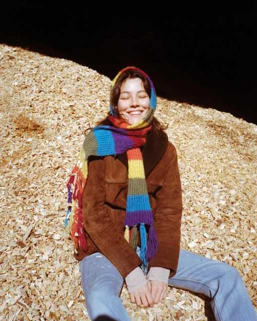 Smiling woman in brown jacket and rainbow scarf sits on wood chips in outdoor light, creating a cheerful and vibrant portrait.