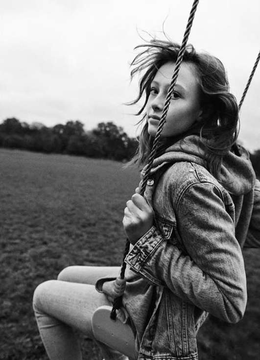 Black and white portrait of a girl on a swing, wearing a denim jacket, with blurred background of field and trees under overcast sky.