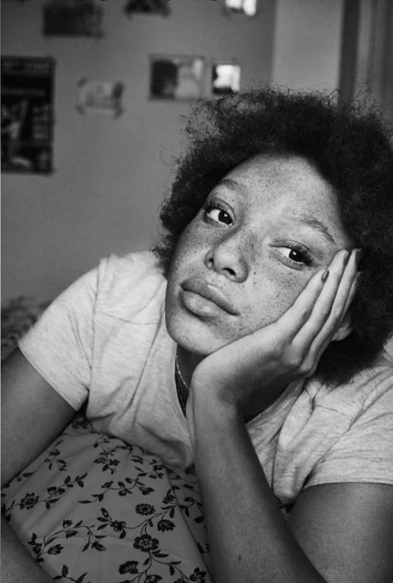 Black and white portrait of a woman with freckles resting her face on her hand, lying on a floral pillow in an indoor setting.