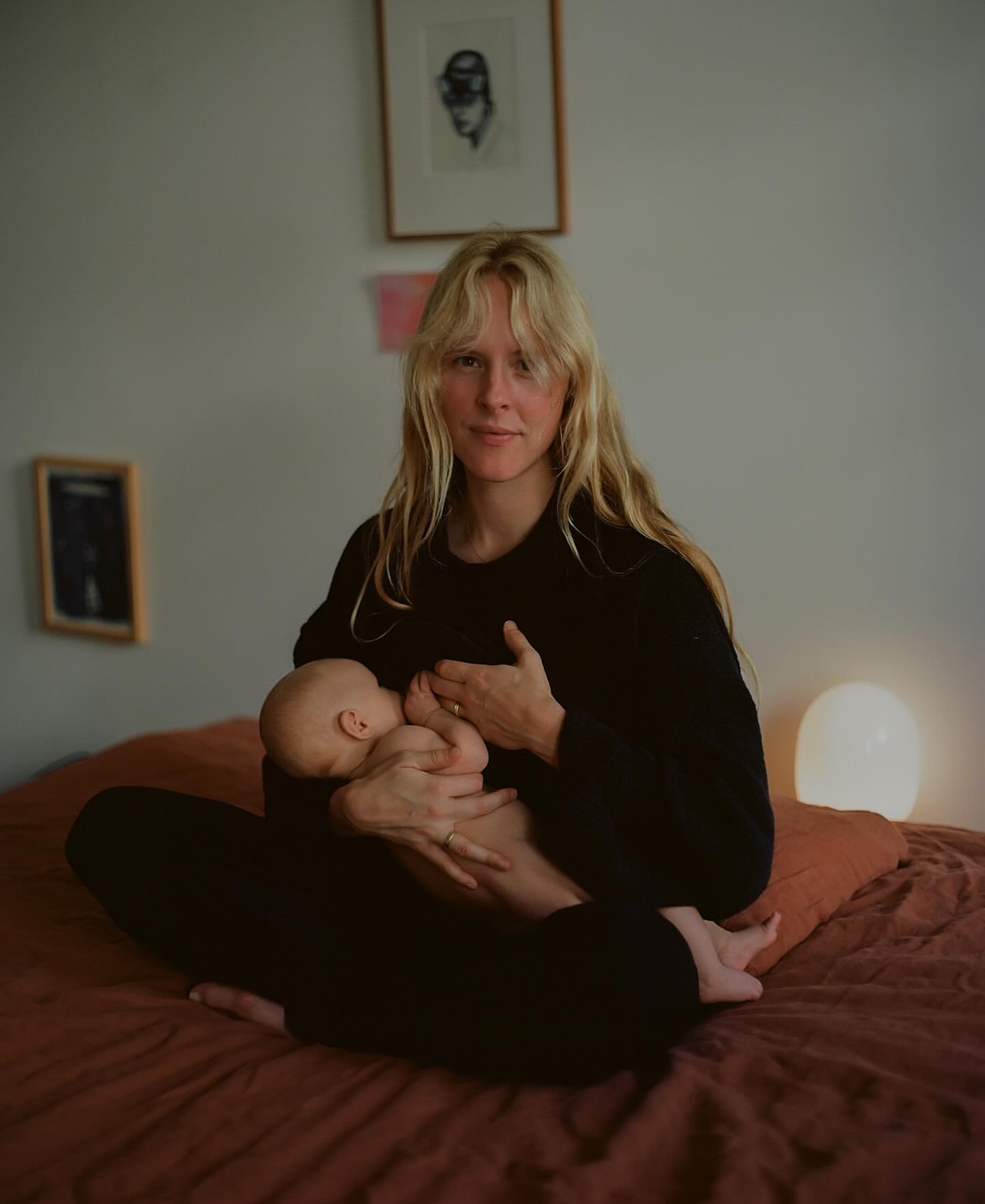 Woman breastfeeding baby indoors. Mother in black top sits cross-legged on terracotta-colored bed with wall art and lamp in background.