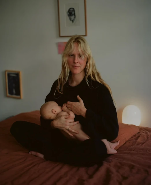 Woman breastfeeding baby indoors. Mother in black top sits cross-legged on terracotta-colored bed with wall art and lamp in background.