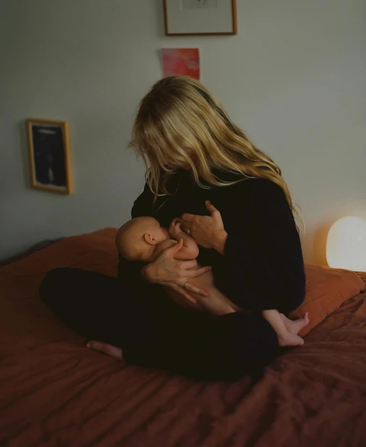 A woman with long blond hair is breastfeeding a baby in a dimly lit bedroom with white walls and a rust-colored bedspread.