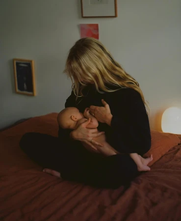 A woman with long blond hair is breastfeeding a baby in a dimly lit bedroom with white walls and a rust-colored bedspread.