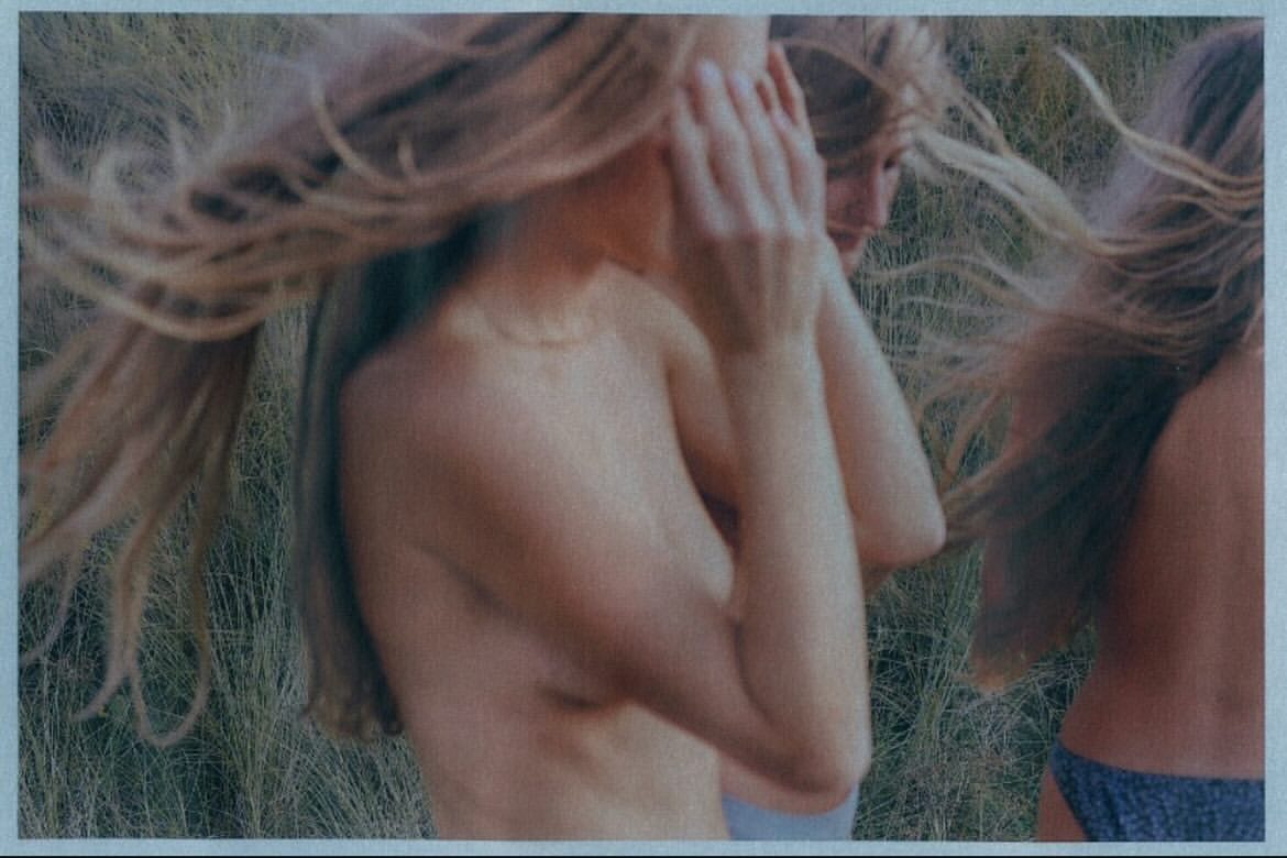 Three fair-skinned women stand outdoors in an artistic photograph featuring blonde hair, bathing suits, and long grass. The focus is soft.