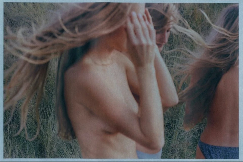 Three fair-skinned women stand outdoors in an artistic photograph featuring blonde hair, bathing suits, and long grass. The focus is soft.