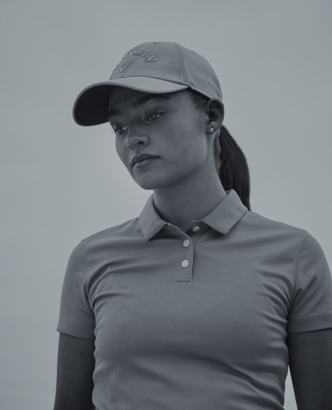Monochrome portrait of a young woman wearing a polo shirt and cap, looking down against a neutral background, in soft studio lighting.