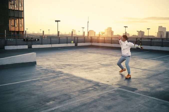 Man in sportswear hits a tennis ball on an outdoor rooftop court with city skyline and soft sunset colors in the sky. 