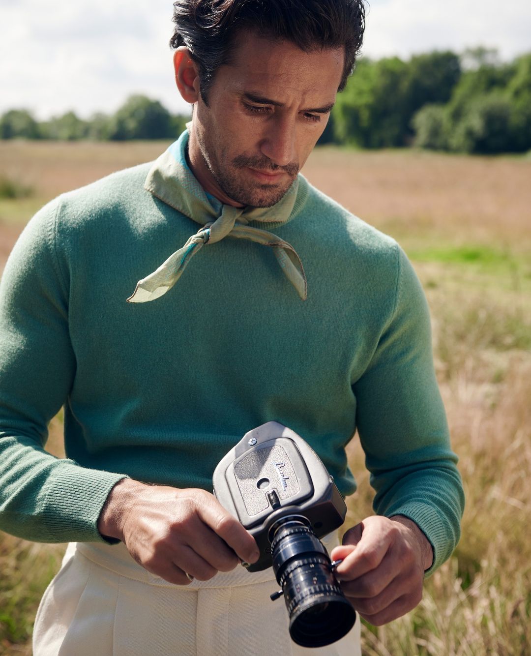 Close-up of a man with dark hair, wearing a green sweater and light pants, holding a vintage film camera in a grassy field outdoors.