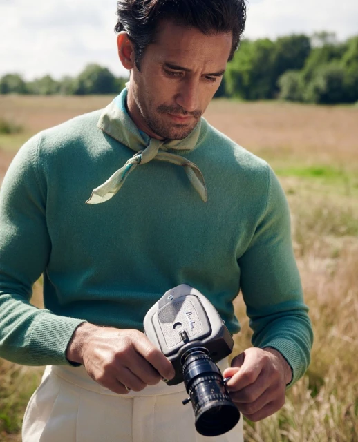 Close-up of a man with dark hair, wearing a green sweater and light pants, holding a vintage film camera in a grassy field outdoors.