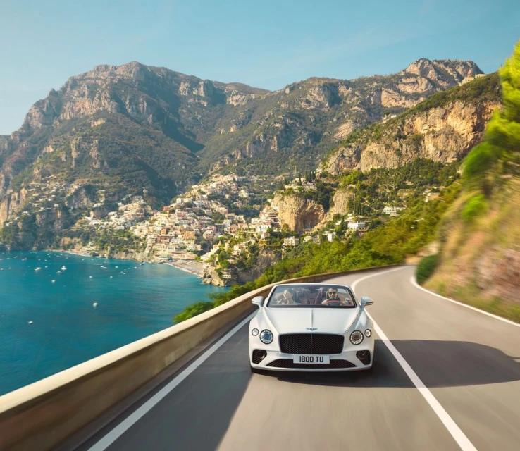White convertible driving on coastal road with mountain and town views, turquoise sea, and clear sky on a sunny day.