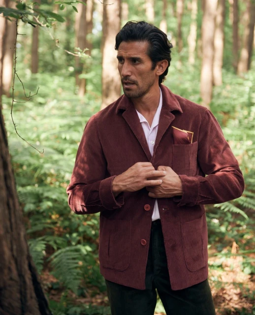 Attractive adult male poses outdoors wearing a maroon jacket and white shirt against a backdrop of lush green foliage and trees.