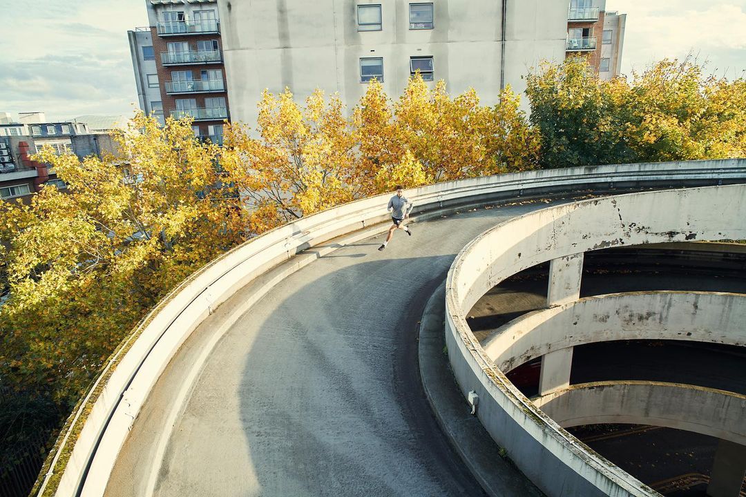 A jogger runs on the spiraling ramp of an urban parking garage, surrounded by trees and city buildings on a cloudy day.