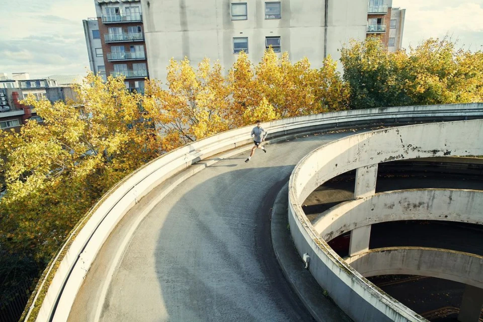 A jogger runs on the spiraling ramp of an urban parking garage, surrounded by trees and city buildings on a cloudy day.