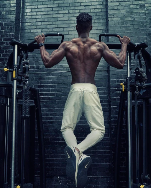 Muscular man in sportswear doing pull-ups at a gym, back view, showcasing strength and fitness in an indoor workout setting.