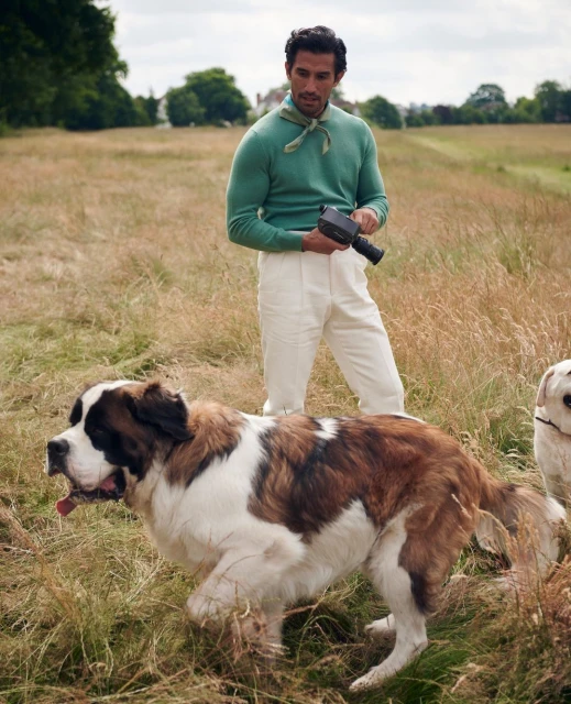 Man in sweater and white pants outdoors with two dogs in a field, one St. Bernard dog and one golden lab, holding vintage camera.