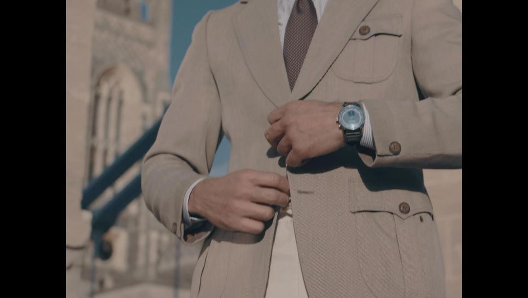 Man in tan suit fastens jacket button in front of an old architectural structure on a sunny day, wearing a watch on his wrist.
