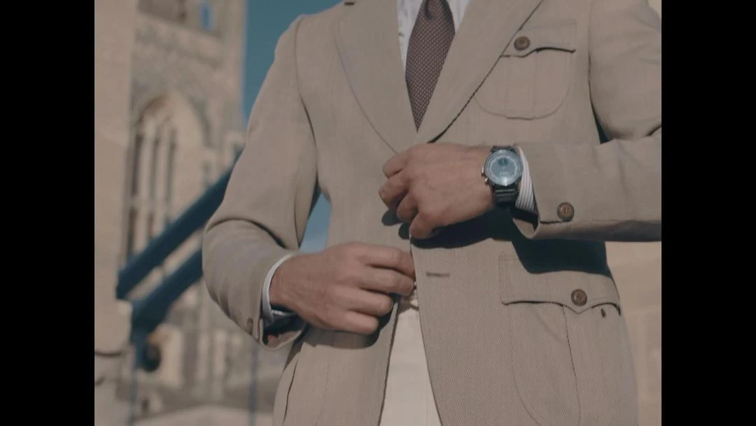 Man in tan suit fastens jacket button in front of an old architectural structure on a sunny day, wearing a watch on his wrist.