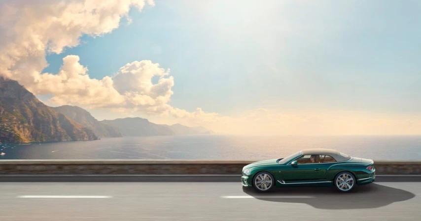 Luxury green convertible driving along a coastal road with mountains, ocean, and a blue sky with clouds in the background.