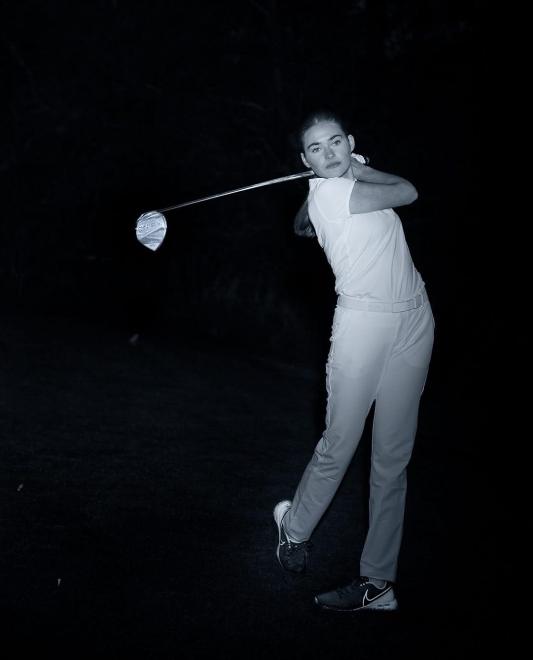 Monochrome shot of a woman swinging a golf club in a dark setting, focusing on her athletic form and the equipment.