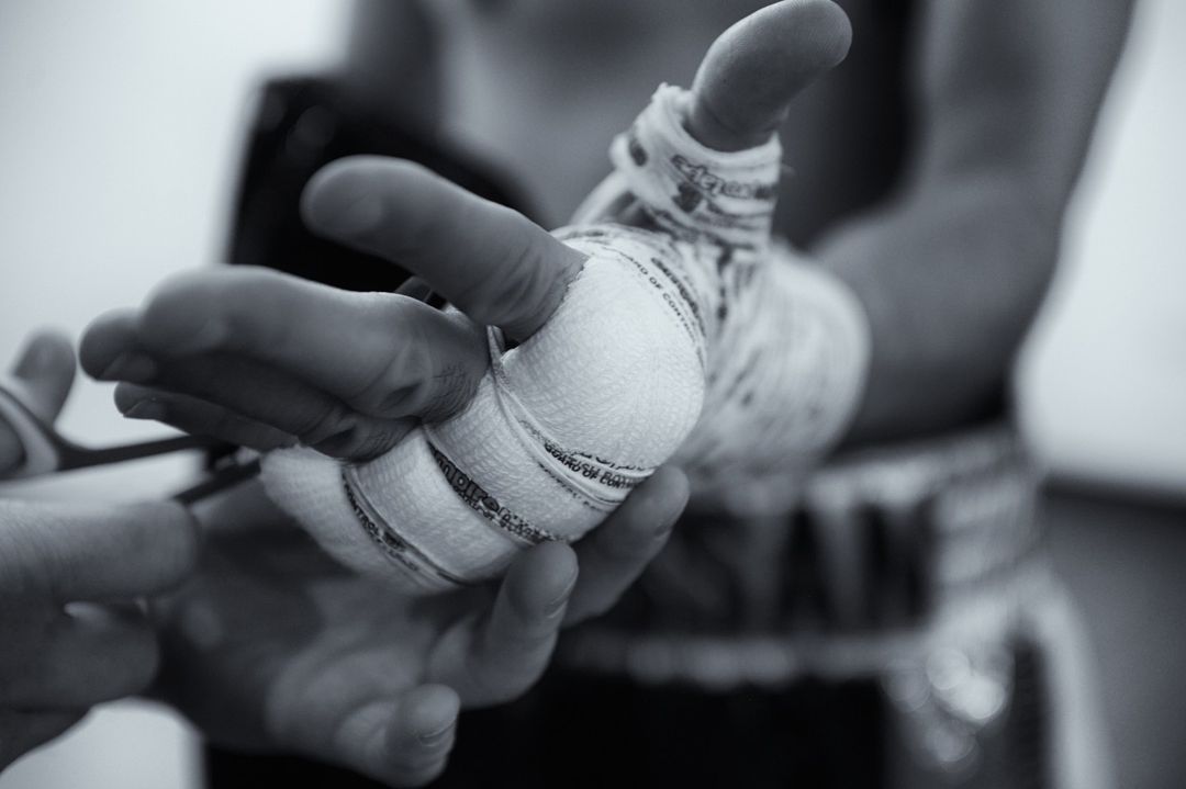 Close-up black and white shot of a person's hand being wrapped in white bandages. Another pair of hands holding scissors is also visible.
