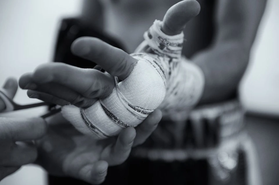 Close-up black and white shot of a person's hand being wrapped in white bandages. Another pair of hands holding scissors is also visible.
