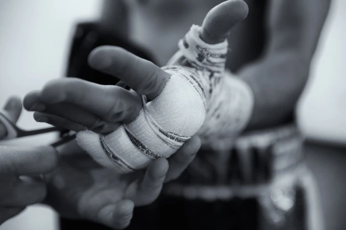 Close-up black and white shot of a person's hand being wrapped in white bandages. Another pair of hands holding scissors is also visible.