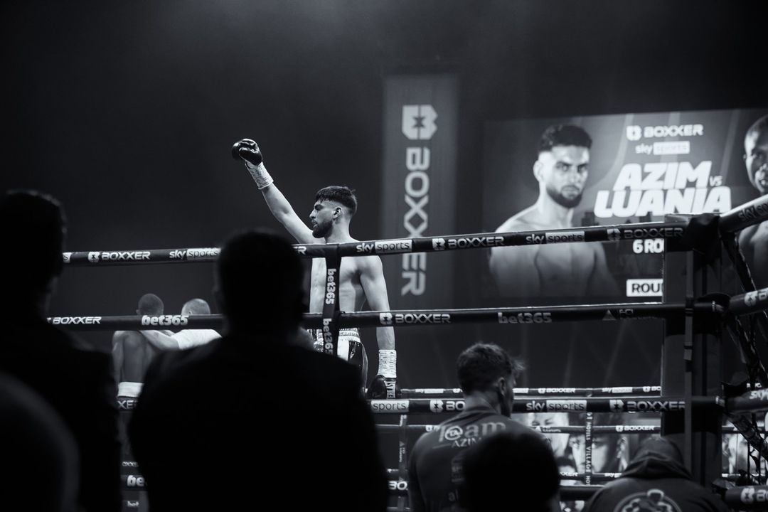 Triumphant boxer raises gloved fist in the ring after winning a match, with spectators and promotional signs in the background, captured in black and white.