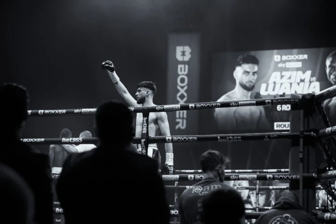 Triumphant boxer raises gloved fist in the ring after winning a match, with spectators and promotional signs in the background, captured in black and white.